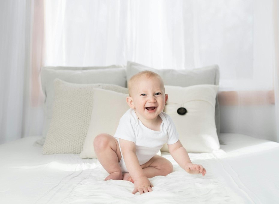 Baby sits on the bed, white tones.